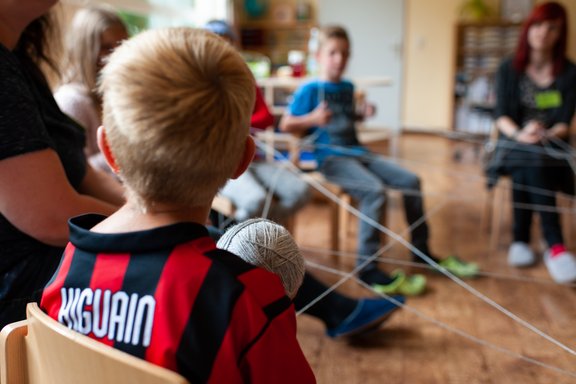 Kinder sitzen auf Stühlen im Kreis und spannen ein Seil.