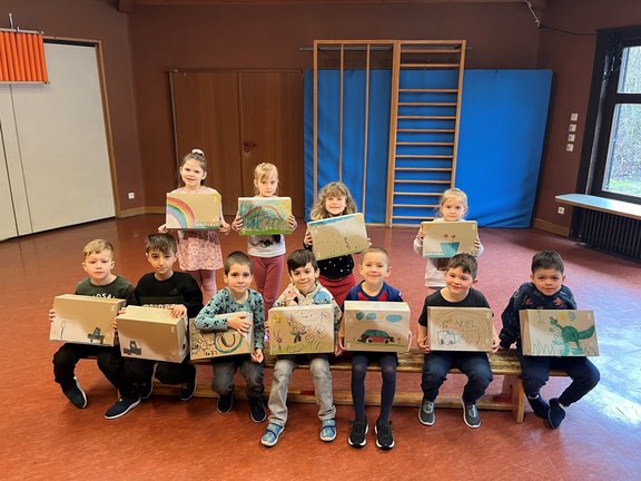 Elf Kinder sind in zwei Reihen aufgestellt und zeigen die Kartons, auf denen bunte Bilder zu sehen sind.