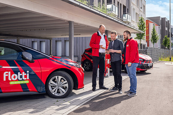 Links steht das Auto in rot lackiert mit dem Schriftzug flotti. Drei Männer stehen zusammen und sprechen. Hr. Gülpen auf der linken Seite erklärt dem Mann in der Mitte etwas am Handy.