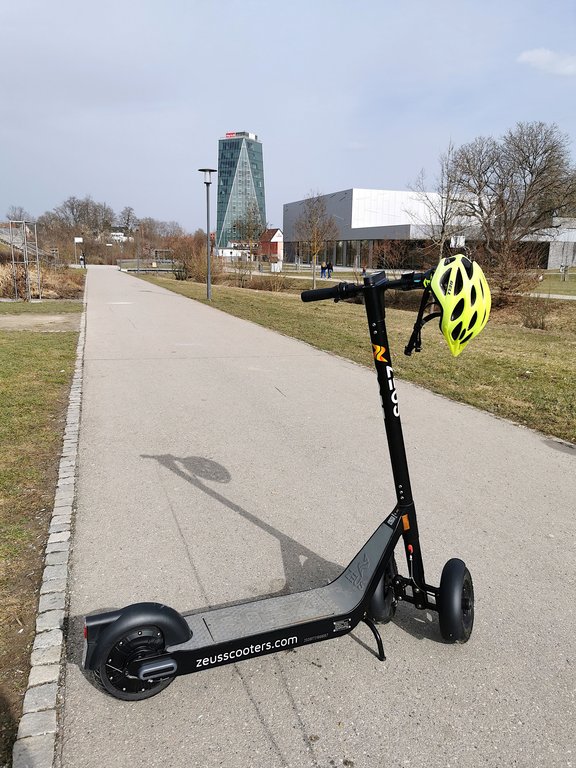 Ein E-Scooter steht auf einem asphaltierten Weg in Schwenningen, der sich durch ein Parkgelände zieht. Im Hintergrund erkennt man einen modernen gläsernen Turm, davor ist eine große Veranstaltungshalle.