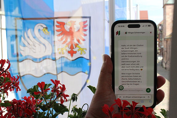 Ein Smartphone zeigt den Chatbot im Display an. Eine Hand hält das Smartphone. Im Hintergrund ist die blau-weiße Stadtfahne mit dem Wappen zu sehen.