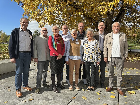 Eine Gruppe von zwölf Personen steht in einem Park mit herbstlichen Bäumen im Hintergrund. Die Menschen lächeln und posieren zusammen, einige tragen farbige Kleidung. Der Boden ist mit herabgefallenen Blättern bedeckt, und der Himmel zeigt sich blau mit einigen Wolken.